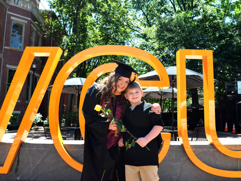 A smiling woman in a cap and gown has an arm over a smiling boy, and they hold a flower together.