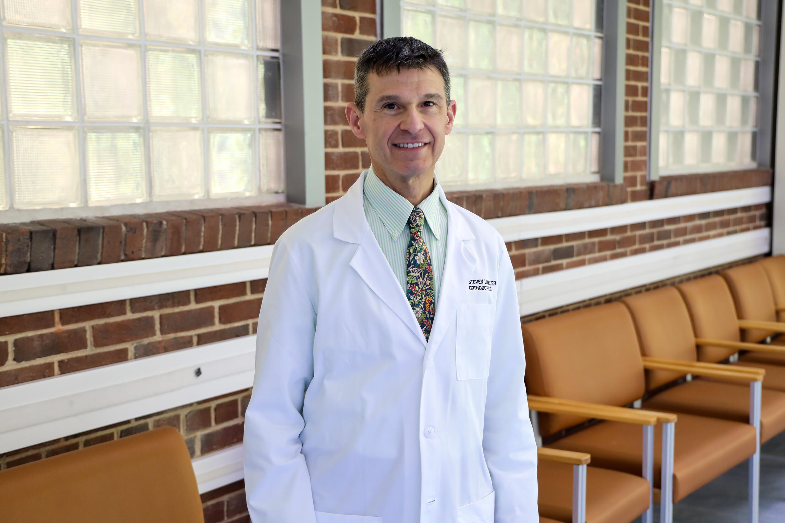 A photo of a man wearing a white dental coat from the waist up. 