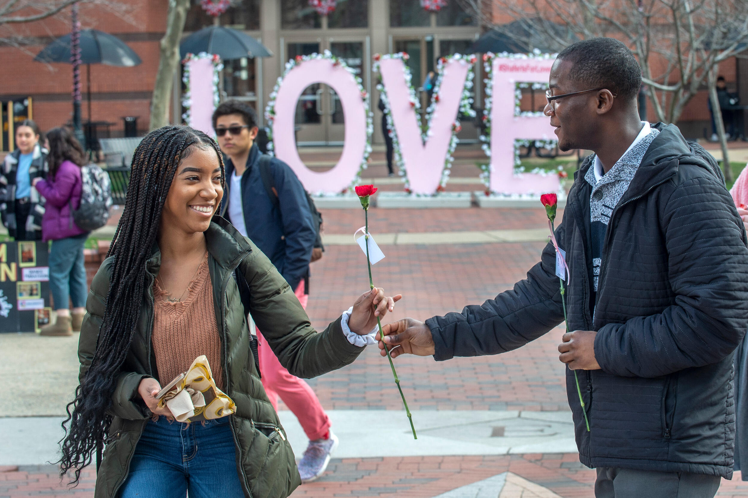 A man holds a flower out to a woman, who is taking it. A large sign spelling out LOVE is in the background.