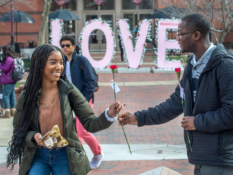 A man holds a flower out to a woman, who is taking it. A large sign spelling out LOVE is in the background.