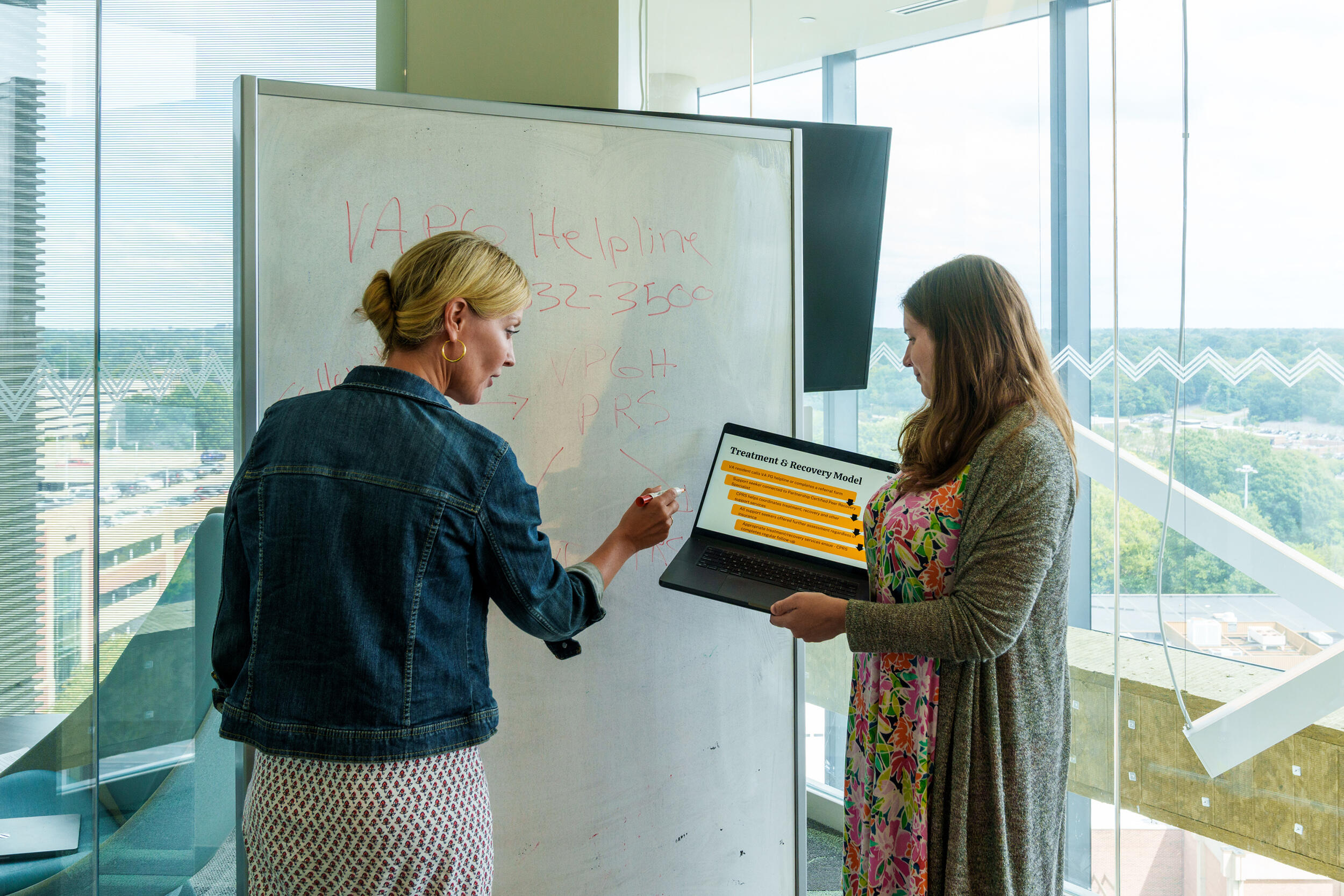 A photo of two women standing in front of a white board. The woman on the left is writing on the board and the woman on the right is holding a laptop that they are both looking at. 