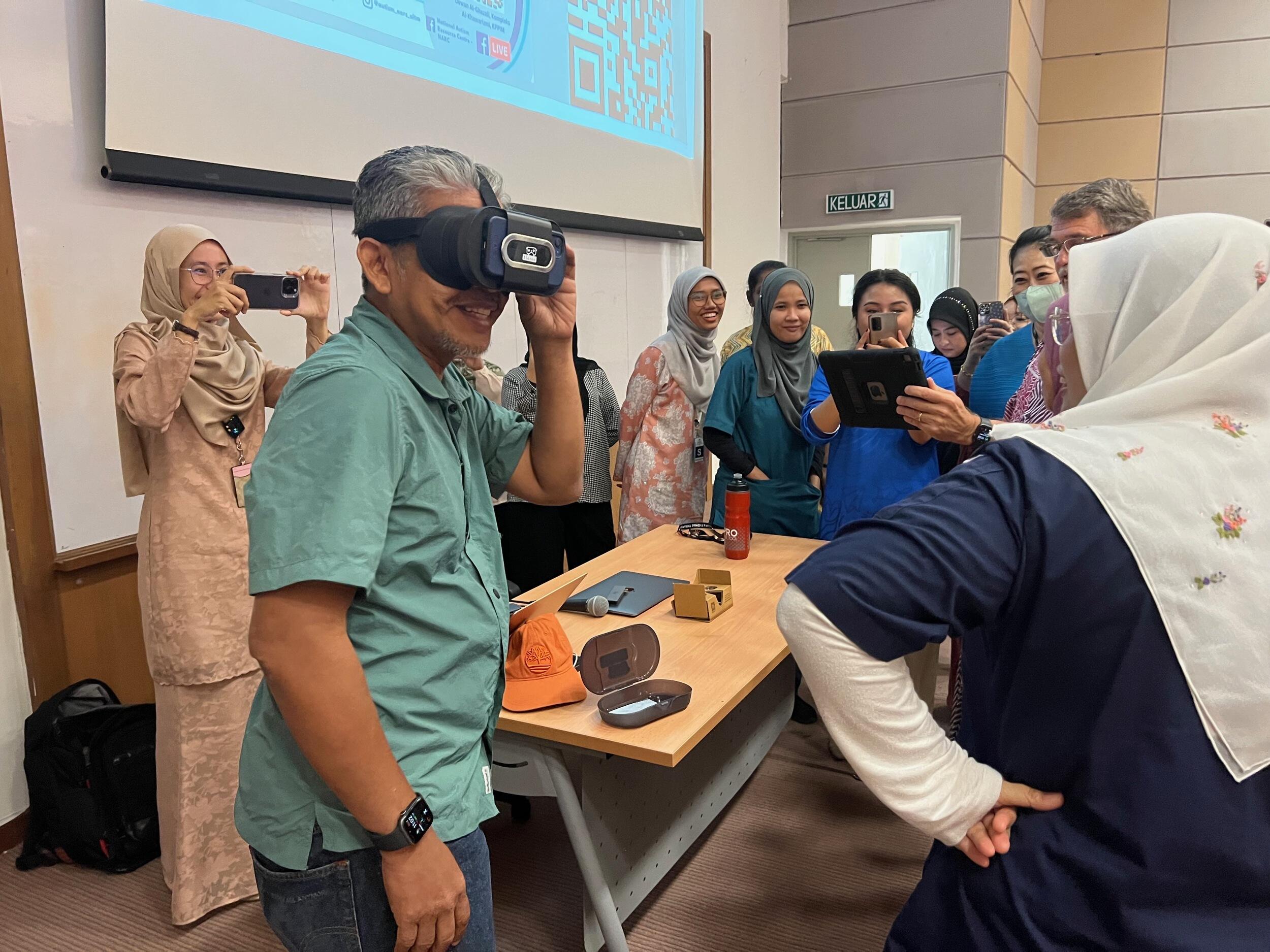 A photo of a man wearing a virtual reality headset in a room full of people. Some of the people in the room are taking photos of him with phones and an ipad. 