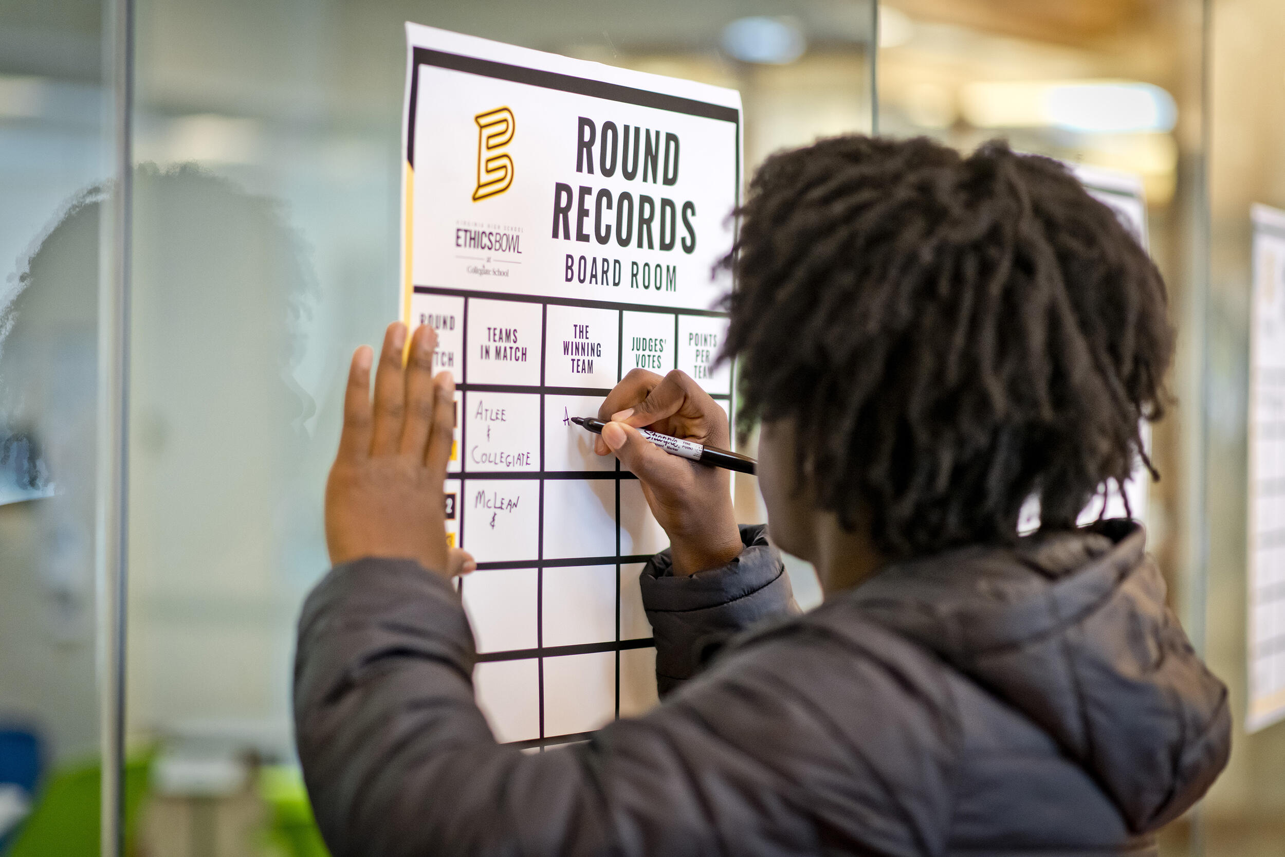 A photo of a teenage boy writing on a piece of paper on a glass window. The top of the paper says \"ROUND RECORDS.\" 