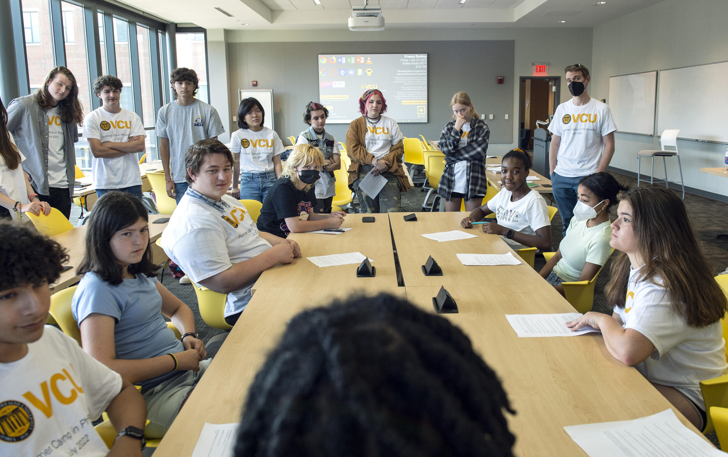 About a dozen students sitting and standing around a long conference room, facing the head of the room.