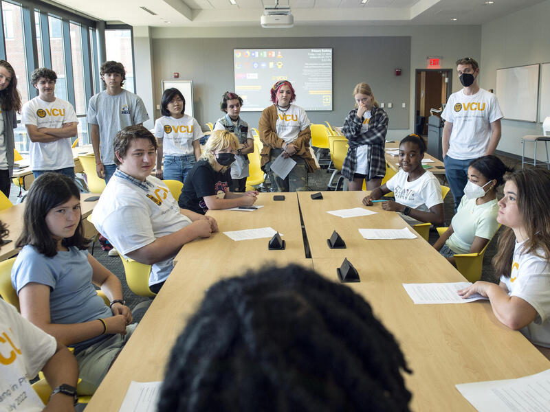 About a dozen students sitting and standing around a long conference room, facing the head of the room.