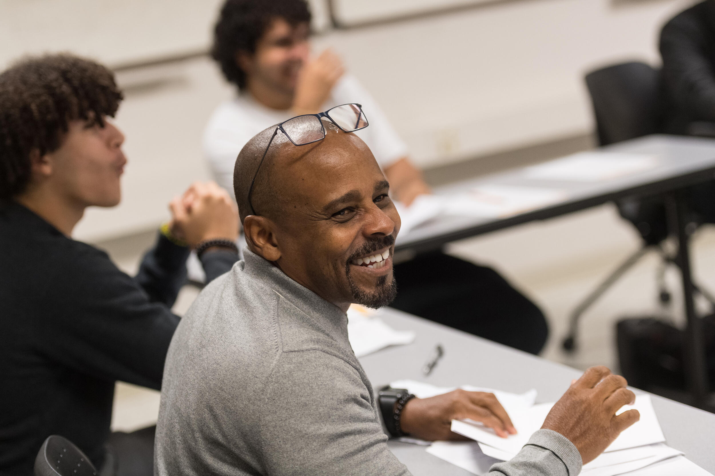 A man smiling and looking left with two younger men sitting to his left.