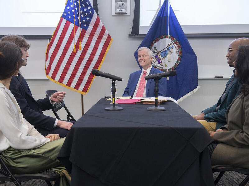 A photo of five people sitting around a rectangle shaped table. Two people sit on the left and right sides of the tame and a man sits at the end of the table. Behind the man is an American flag and a Virginia state flag. 