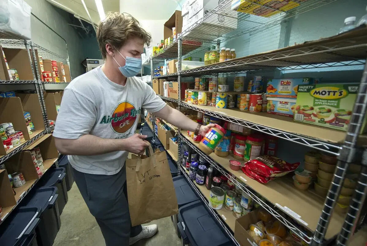 A person wearing a face mask putting a jar of peanut butter on a shelf filled with food 