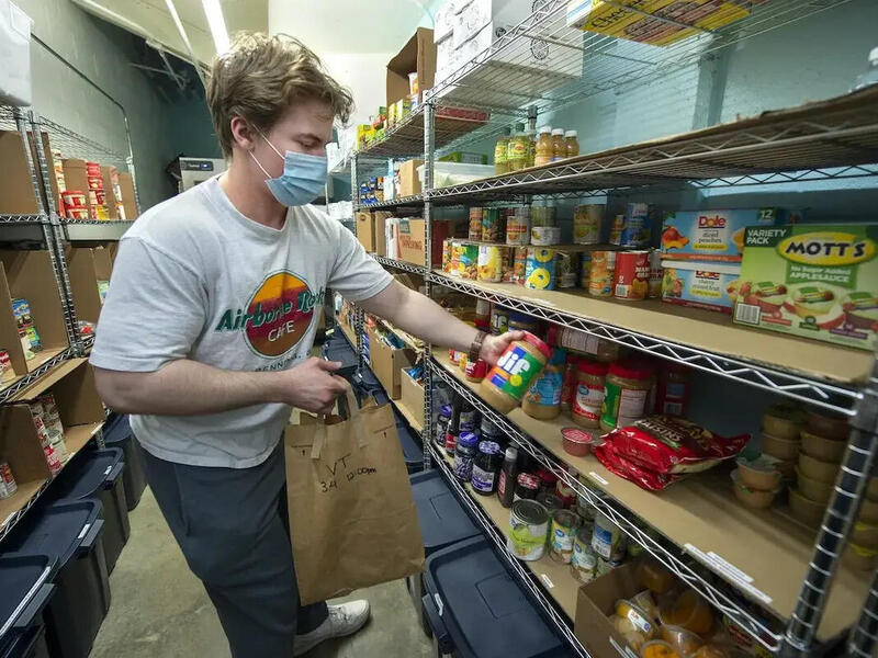 A person wearing a face mask putting a jar of peanut butter on a shelf filled with food 