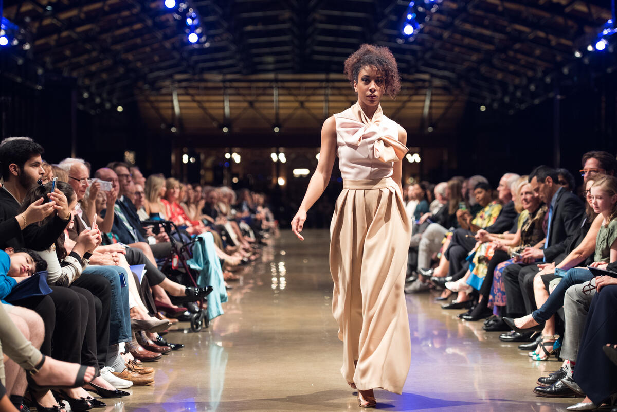 A woman in a full length beige dressing walking down a runway with a line of people sitting on her left and right