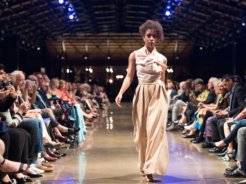 A woman in a full length beige dressing walking down a runway with a line of people sitting on her left and right