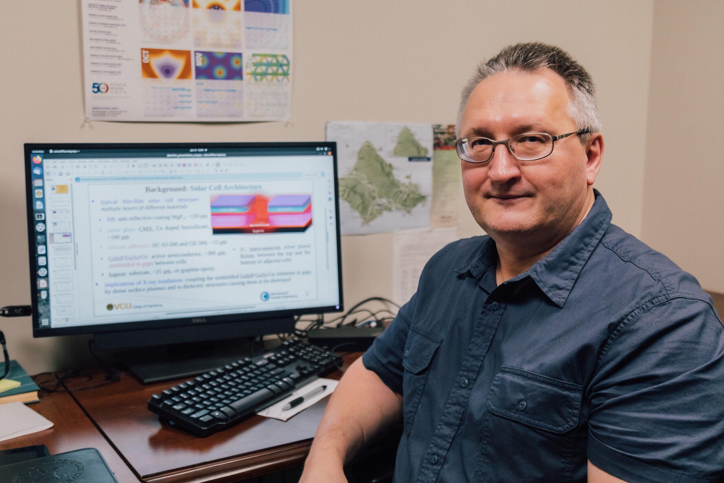 A man wearing a blue button up shirt and classes facing the gamer. He is sitting at a desk with a computer monitor and keyboard on it. 