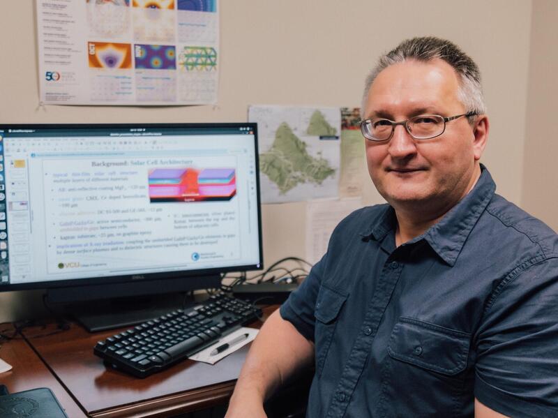 A man wearing a blue button up shirt and classes facing the gamer. He is sitting at a desk with a computer monitor and keyboard on it. 