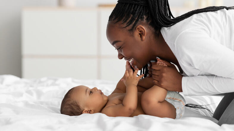 A photo of a woman lenaing over a baby and smilining. She is holding the baby's legs while the baby is lying down, and the baby is touching the woman's face. 
