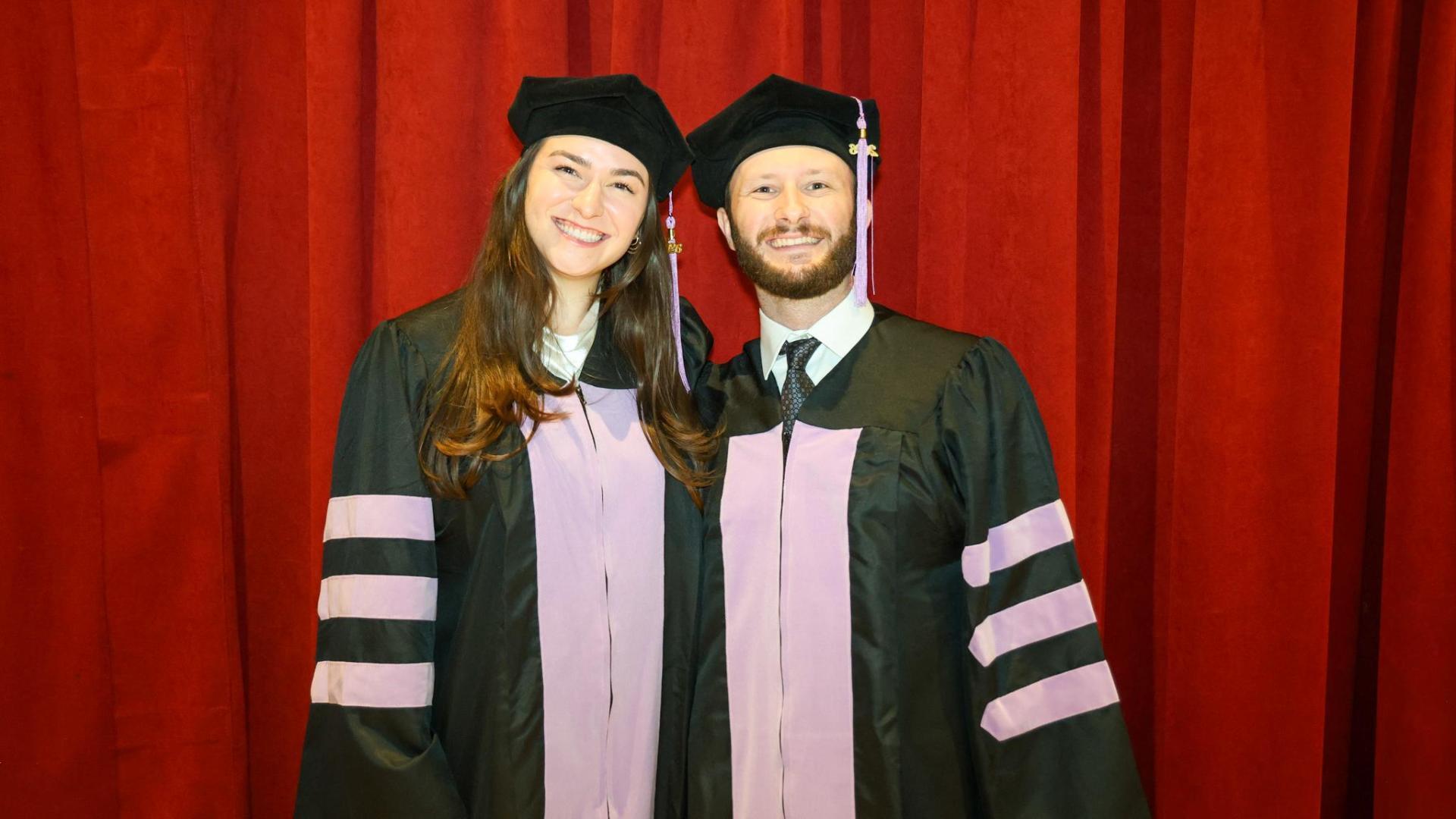 A photo of a man and woman wearing graduation cap and gowns standing next to eachother in front of a red curtain. 