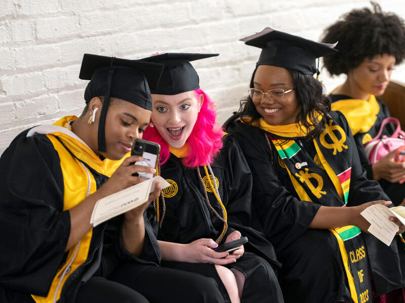 A photo of four students wearing graduation caps and gowns sitting in a row. 