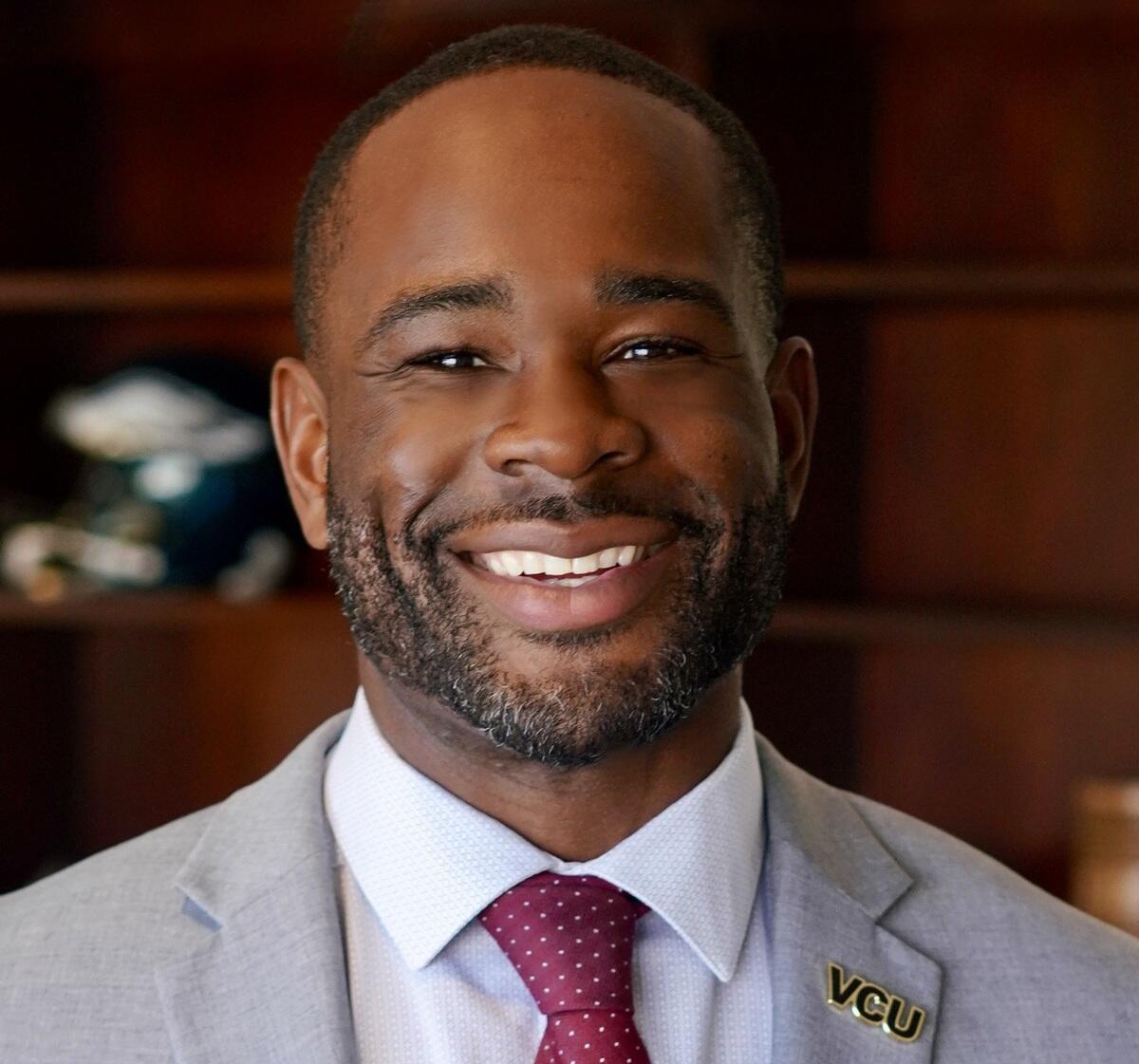 A man wearing a suit and tie smiling. On the collar of his sports coat is a pin that says \"VCU\"