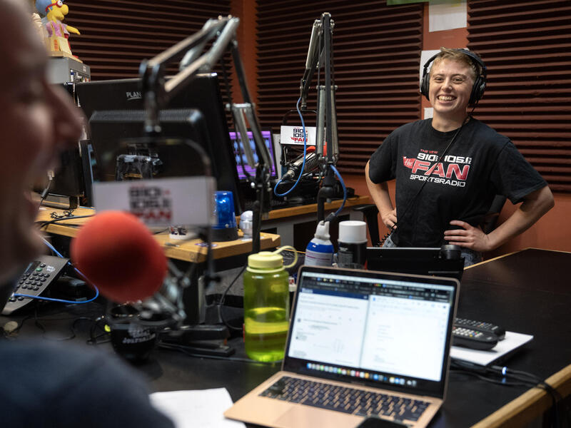 A photo of a man from the waist up smiling on the side of a radio production room. He is wearing headphones and his hands are on his hips. In the foreground is a man from the shoulders up who is smiling. The room is filled with microphones, tables, and computer equipment sitting on the tables.