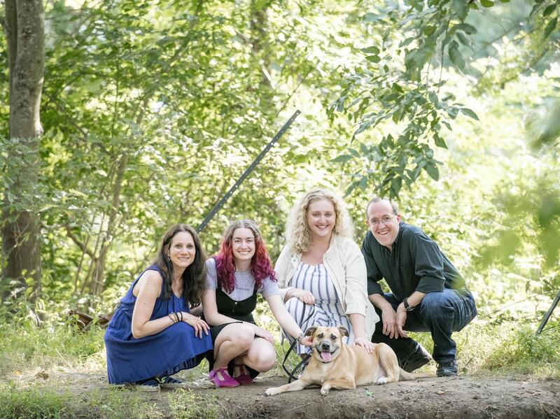 A photo of four people crouching in the woods behind a dog laying on the ground. 