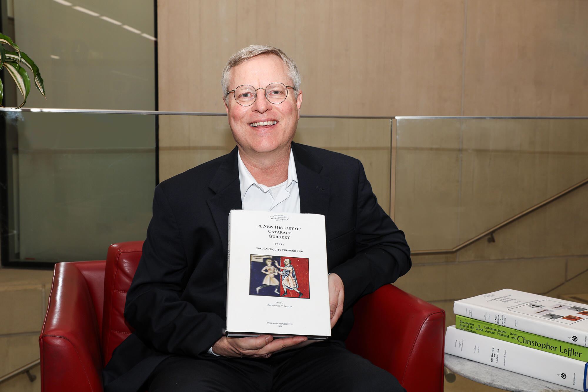 A photo of a man sitting in a red chair. He is holding a book and smiling. 
