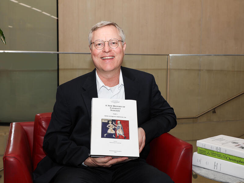 A photo of a man sitting in a red chair. He is holding a book and smiling. 