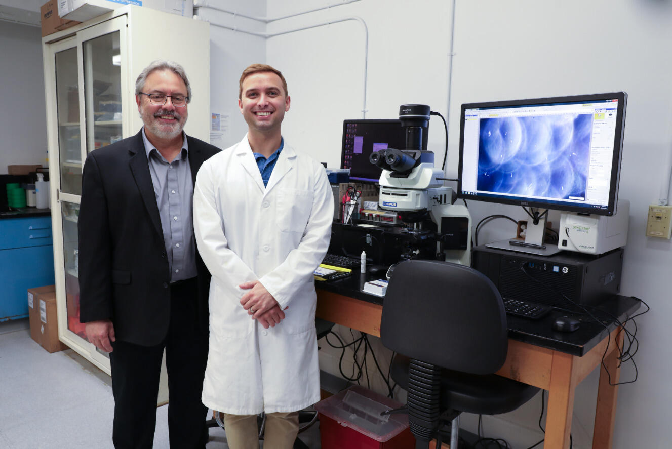 A photo of two men standing next to a table with a magnifying class hooked up to a computer and monitor. The computer monitor shows an image with blue and white circles on it.
