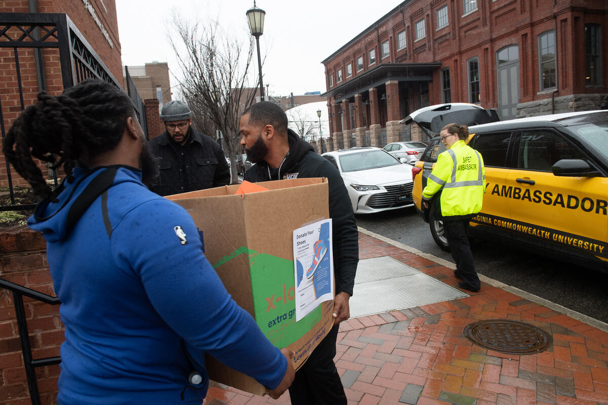 A photo of two men carying a box out on a sidewalk. A man on the left is looking at them and another person on the right is standing next to an SUV with an open trunk. 