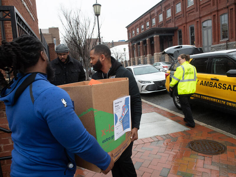 A photo of two men carying a box out on a sidewalk. A man on the left is looking at them and another person on the right is standing next to an SUV with an open trunk. 