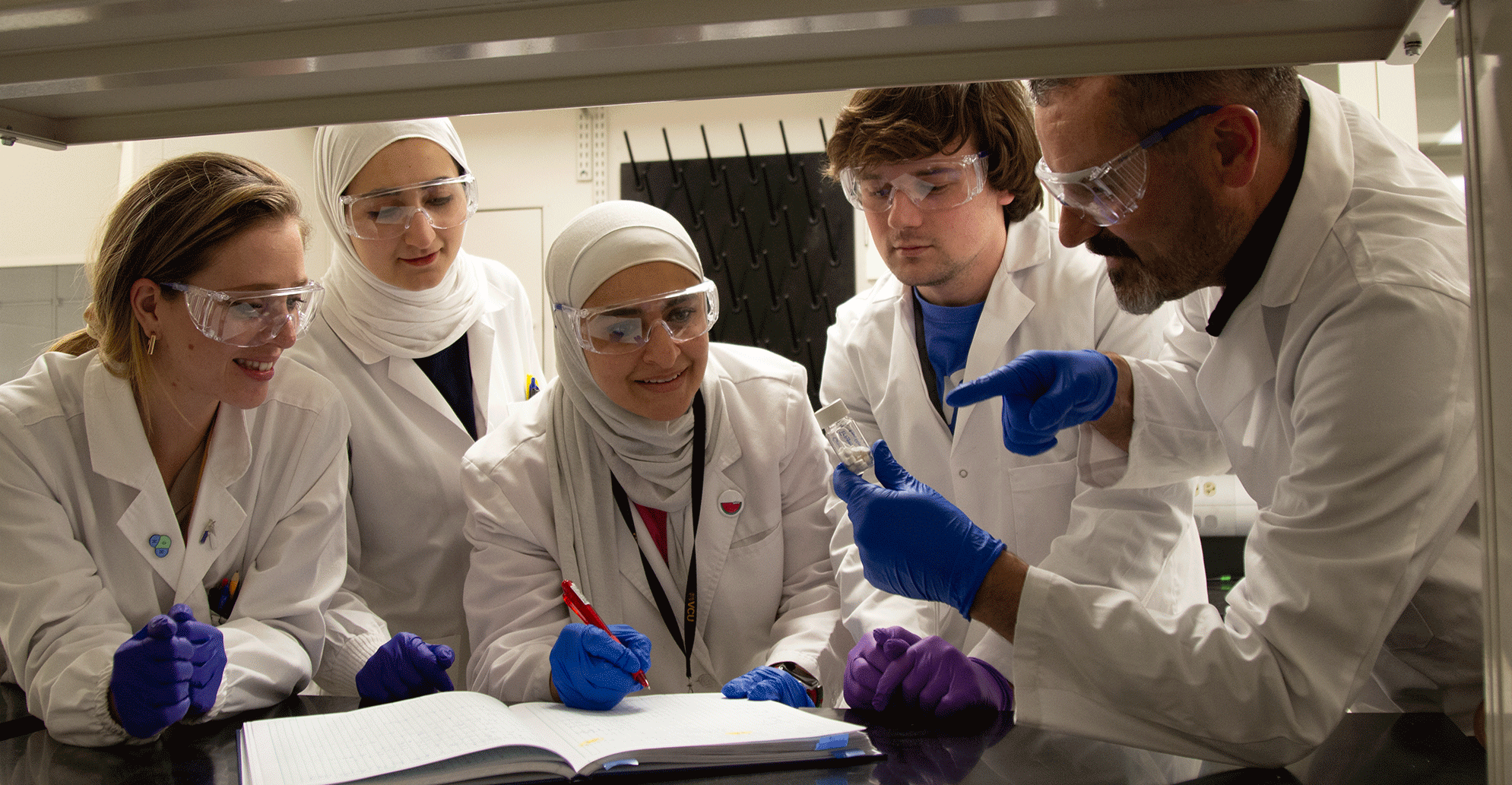 A photo of five people wearing white lab coats, clear safety glasses, and blue latex gloves standing around a table. 