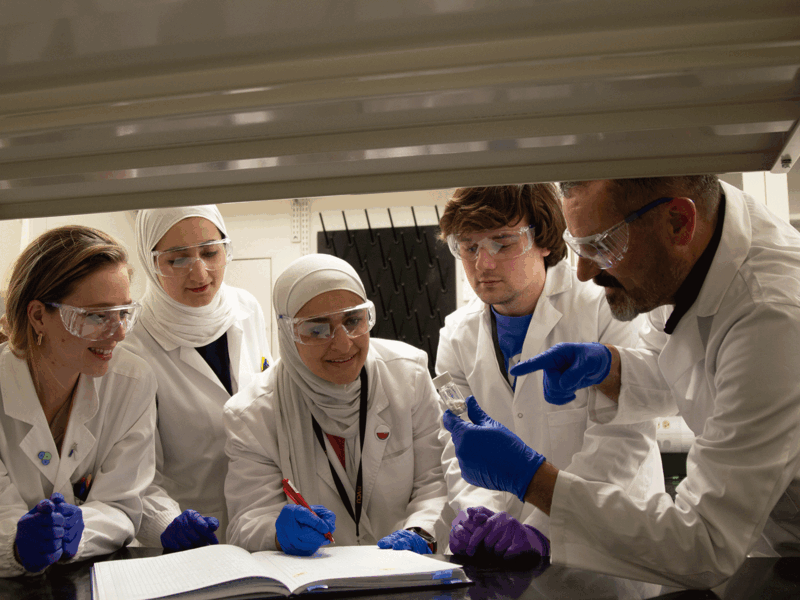 A photo of five people wearing white lab coats, clear safety glasses, and blue latex gloves standing around a table. 