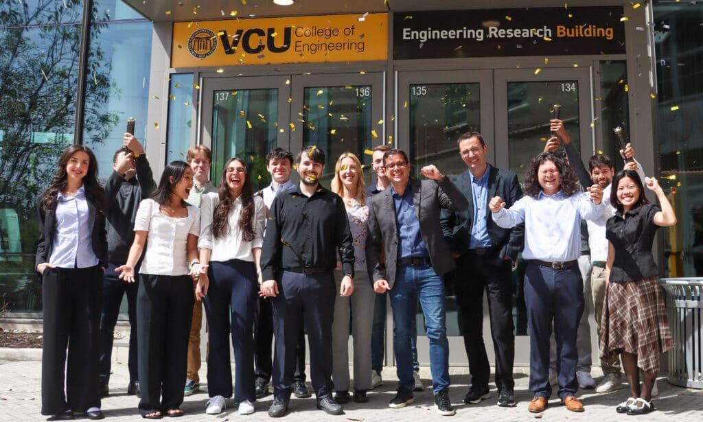 A group photo of 15 people. Two people in the back of the group are shooting confetti. Behind are the front doors to a building. Above the doors are signs that read \"VCU College of Engineering\" \"Engineering Research Building.\" 