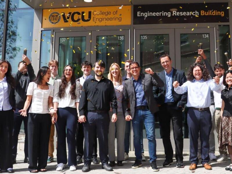 A group photo of 15 people. Two people in the back of the group are shooting confetti. Behind are the front doors to a building. Above the doors are signs that read \"VCU College of Engineering\" \"Engineering Research Building.\" 