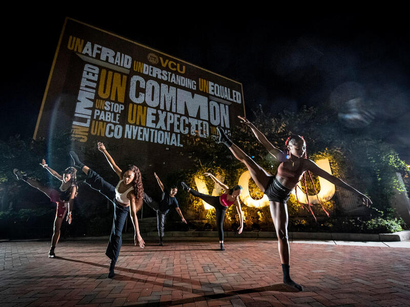 Students participate in a dance performance at night under a wall lit with an artistic word cloud of words that fit with the uncommon experience VCU provides: Unafraid, unlimited, understanding, unequaled, uncommon, unexpected, unstoppable and unconventional.