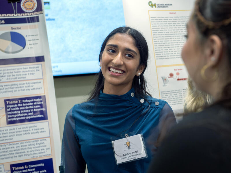 This is a photo of a woman smiling at another woman facing her. The first woman is standing to the right of a poster with research on it. 