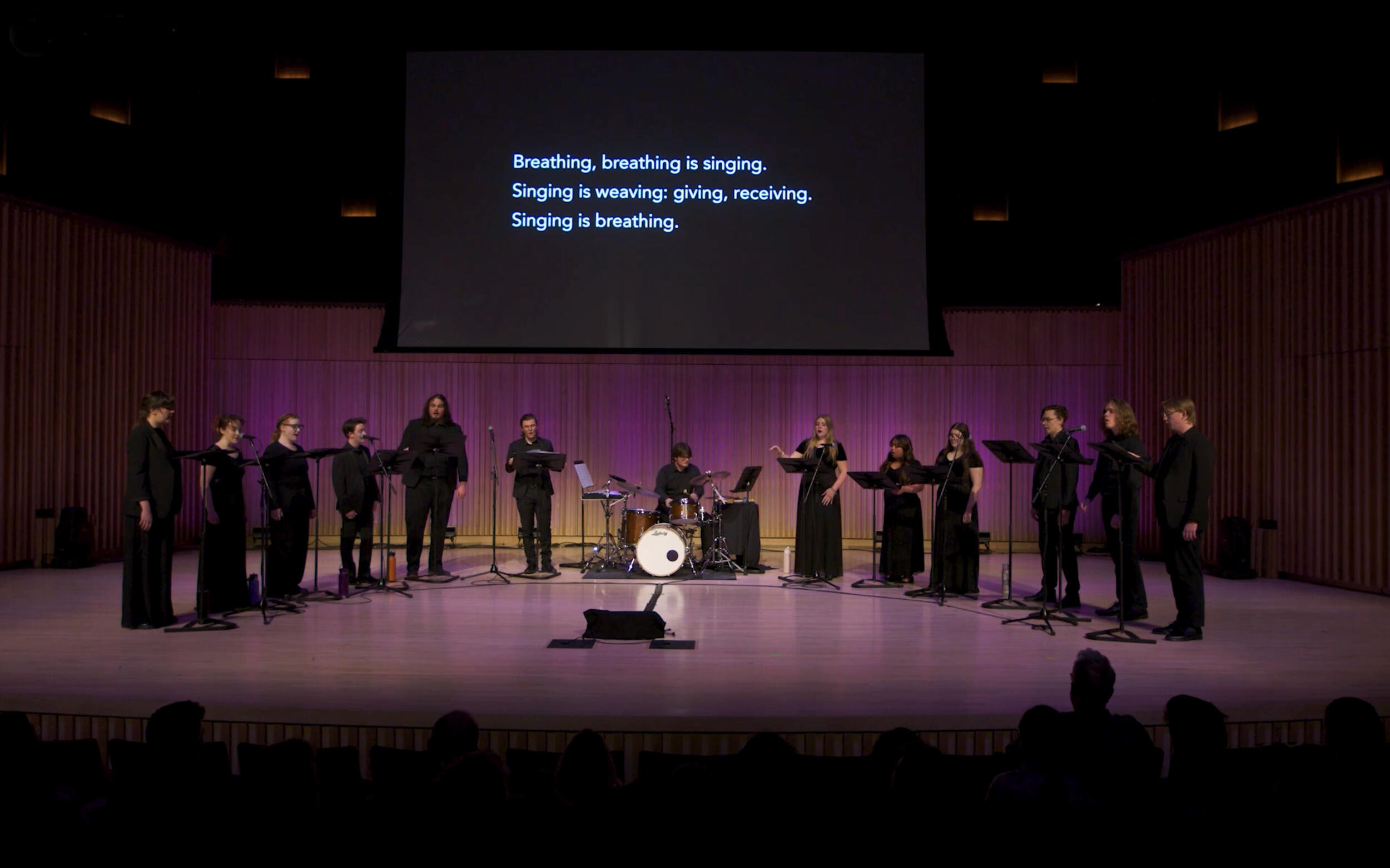 A photo of 11 singers on a stage with a drummer at a drum set sitting in the middle of the group. A black screen behind them has white text that reads \"Breathing, breathing is sining.\" \"Singing is giving: weaving and receiving.\" \"Singing is breathing.\"