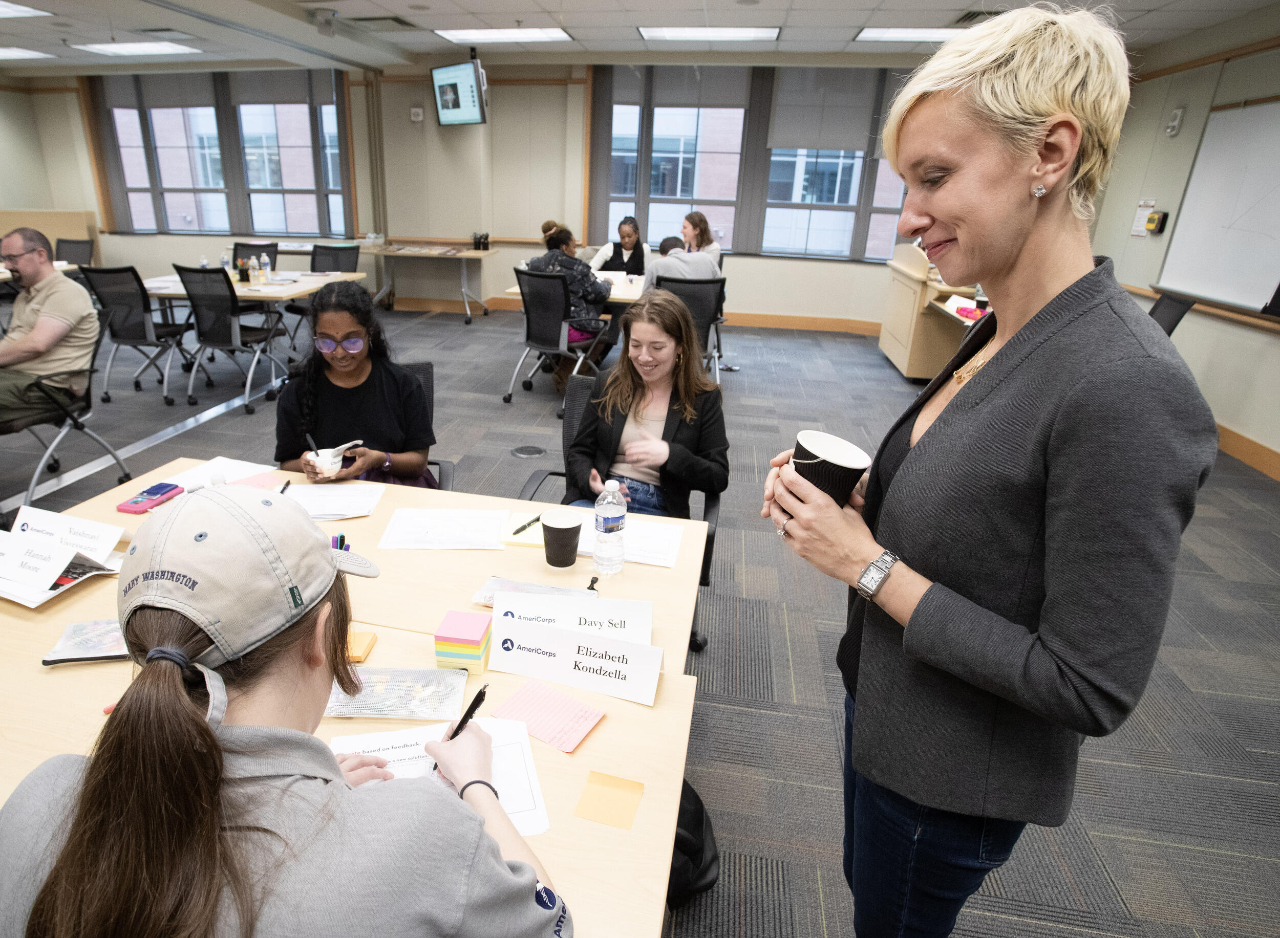 A photo of a woman holding a paper cup smiling while standing next to a table with three students sitting at it. 