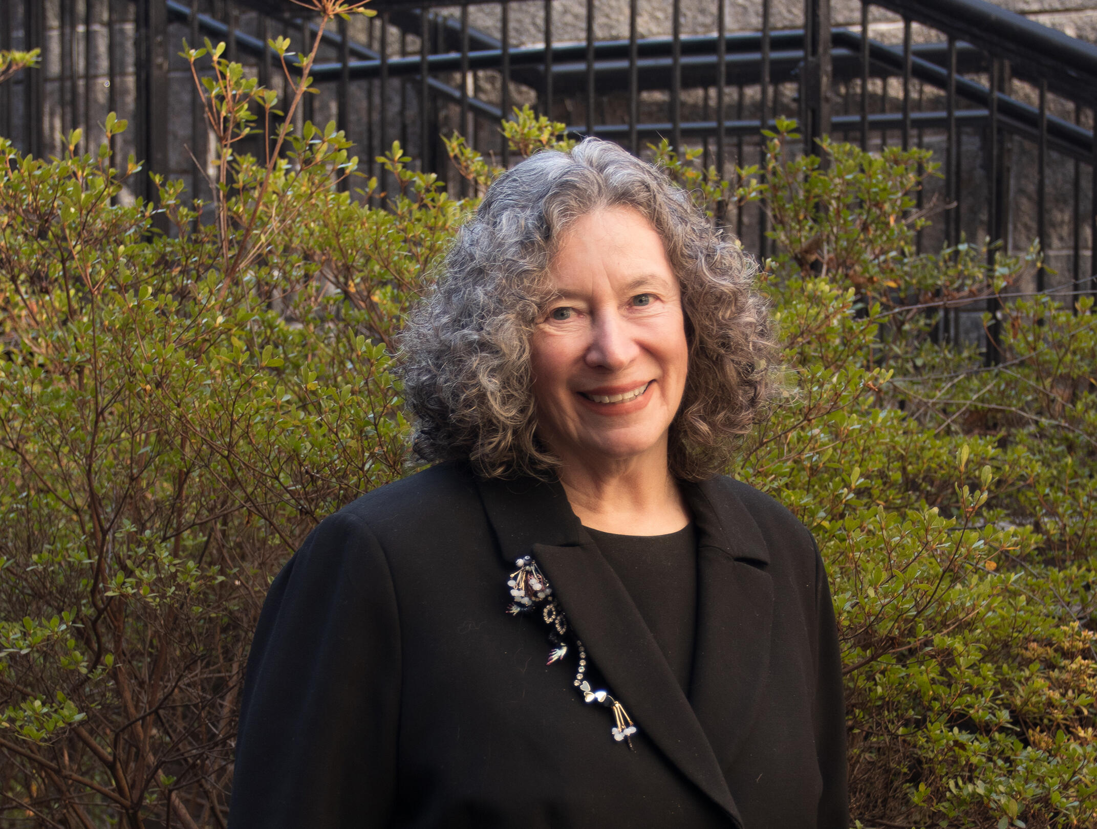 A photo of a woman from the chest up. She is standing in front of some bushes. 