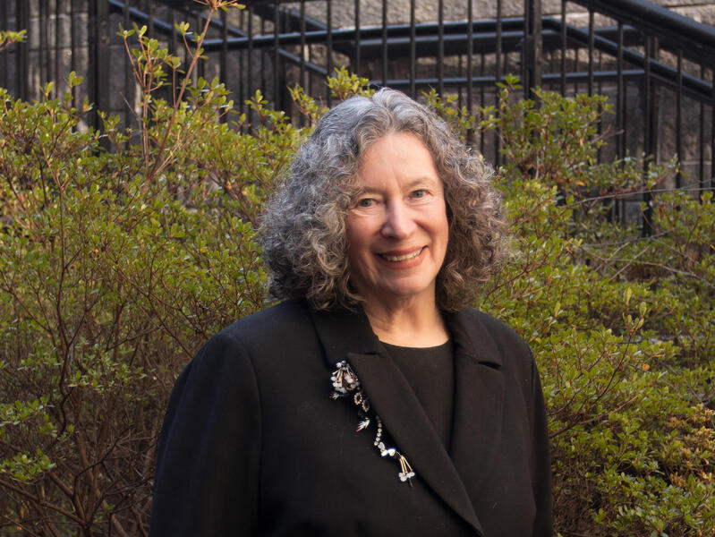 A photo of a woman from the chest up. She is standing in front of some bushes. 