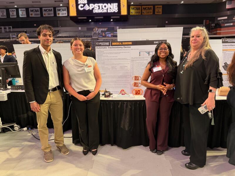 A photo of four people standing in front of a table with a research poster on it. 