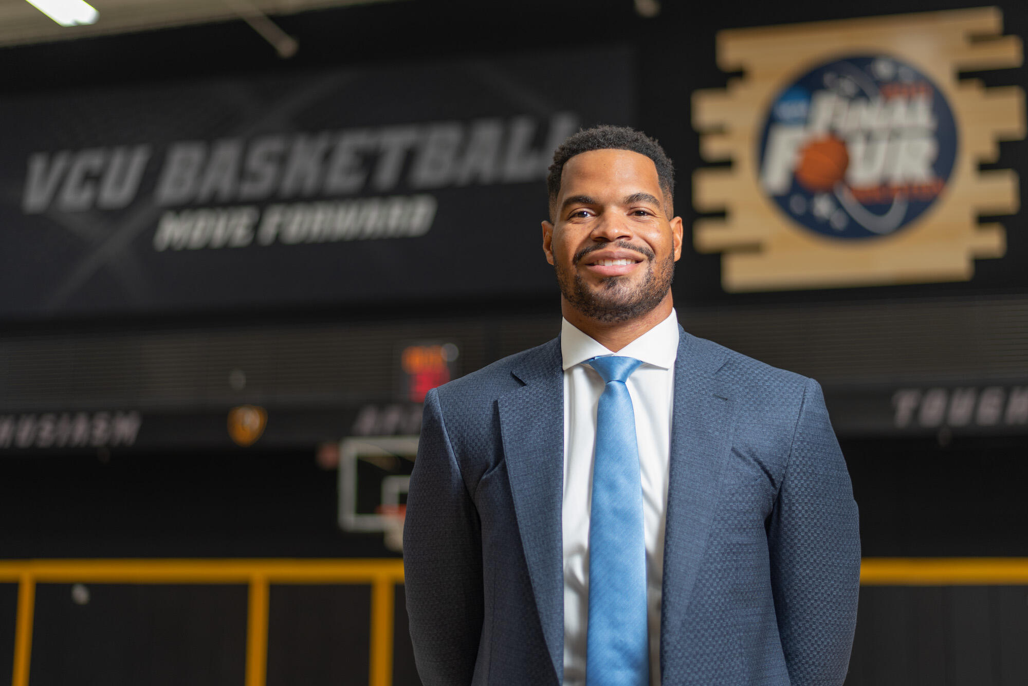 Smiling man in coat and tie standing in front of VCU Basketball sign.