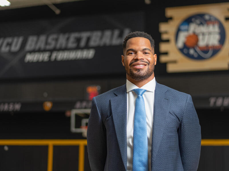 Smiling man in coat and tie standing in front of VCU Basketball sign.