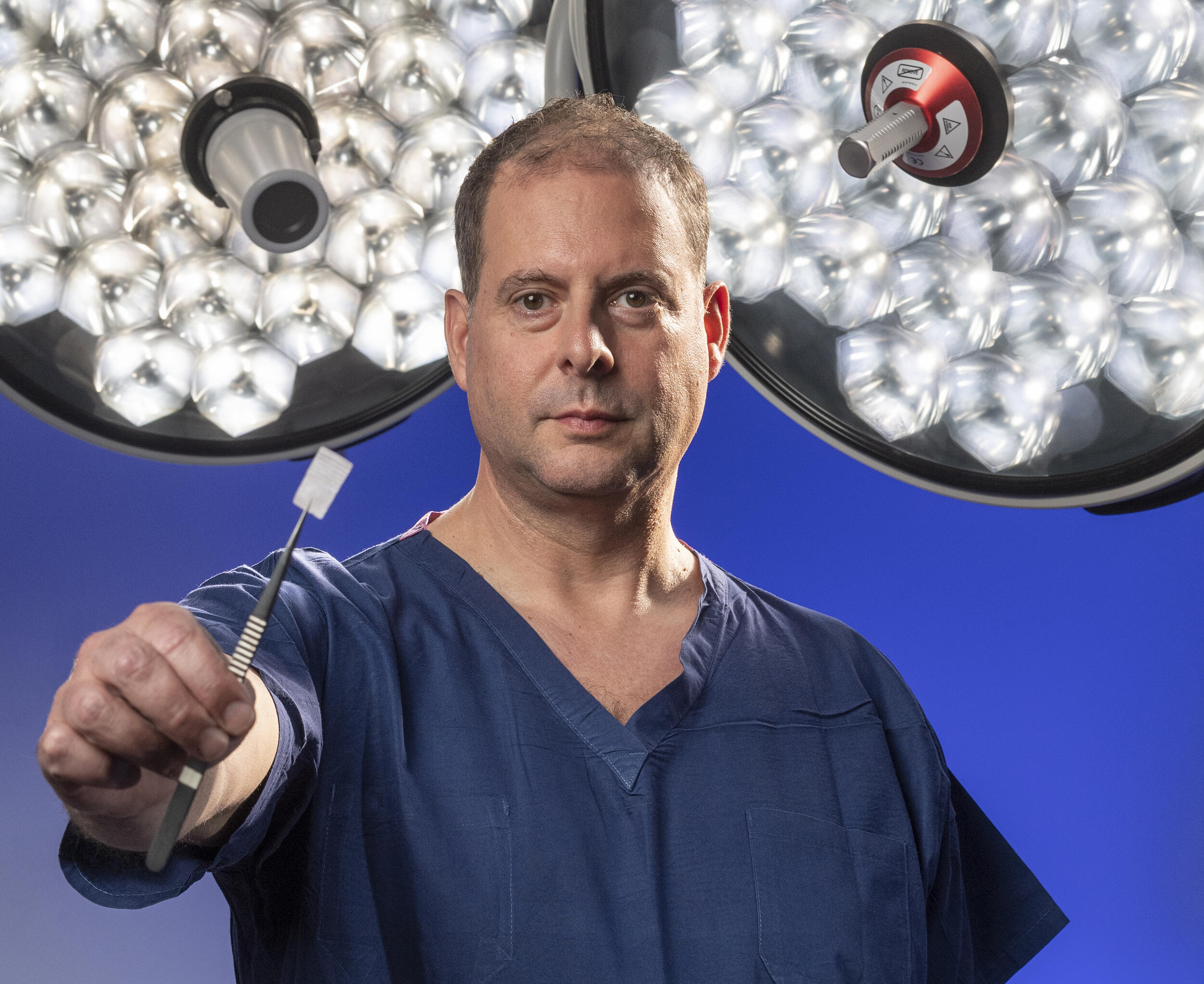 A man in blue scrubs holds up a small rectangular piece of some kind of tape with lights behind him.