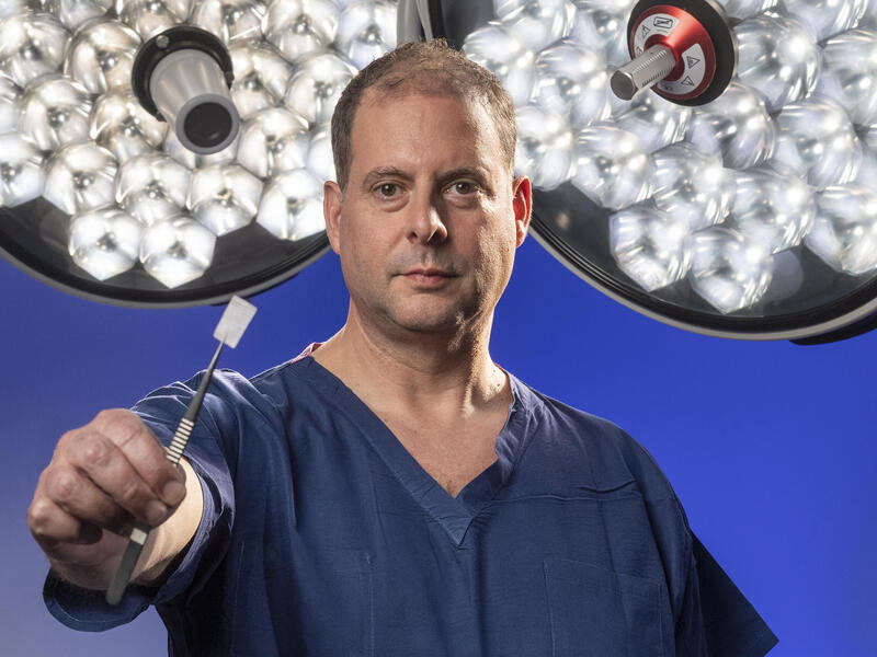 A man in blue scrubs holds up a small rectangular piece of some kind of tape with lights behind him.