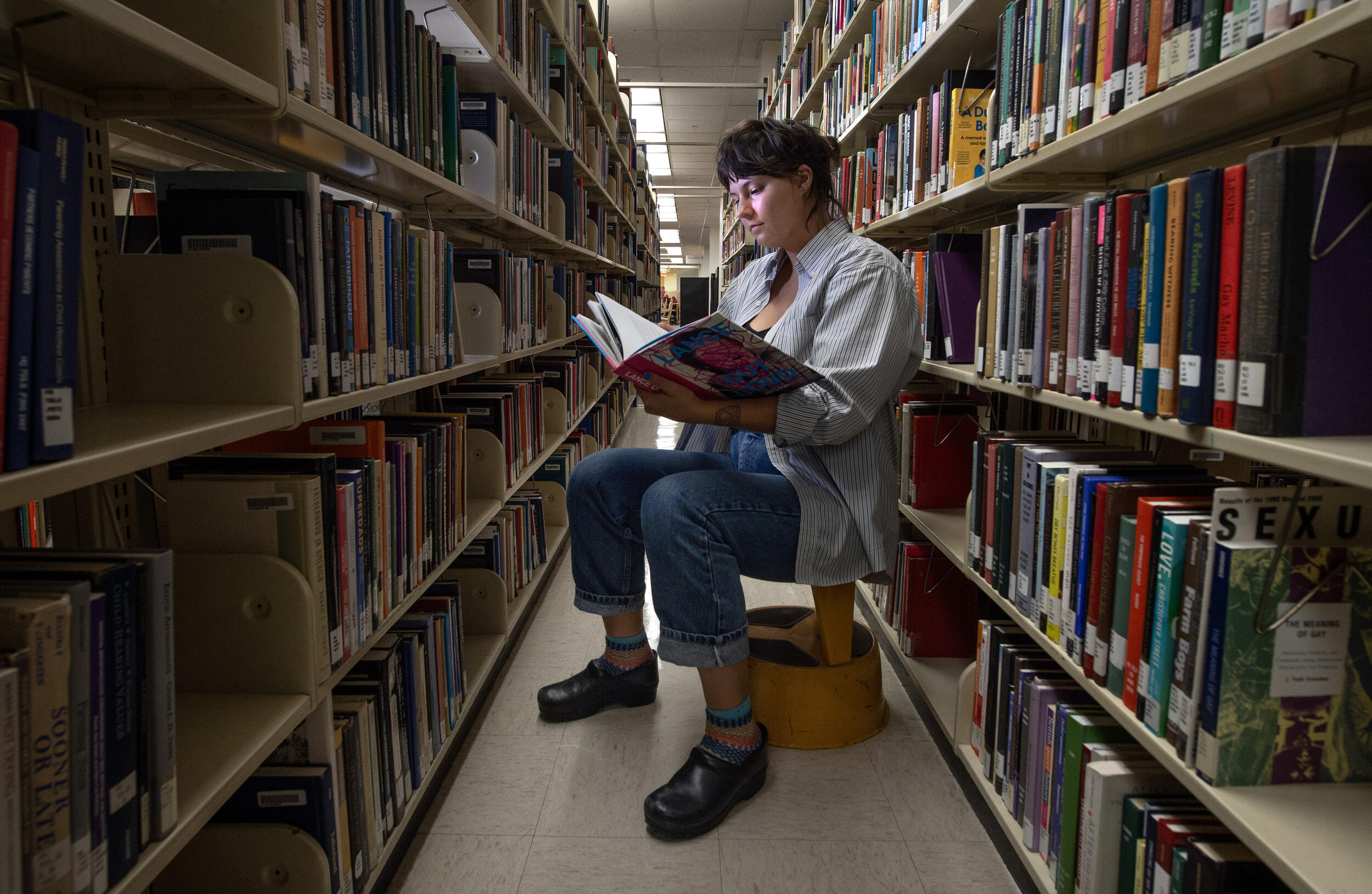 A photo of a woman siting on a stool in between two rows of books in a library. She is reading a book that she is holding with her right hand. 