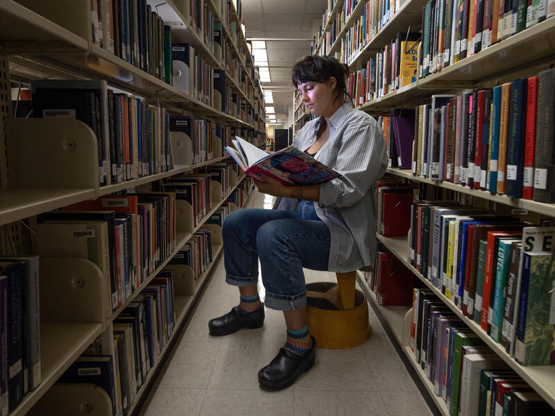 A photo of a woman siting on a stool in between two rows of books in a library. She is reading a book that she is holding with her right hand. 