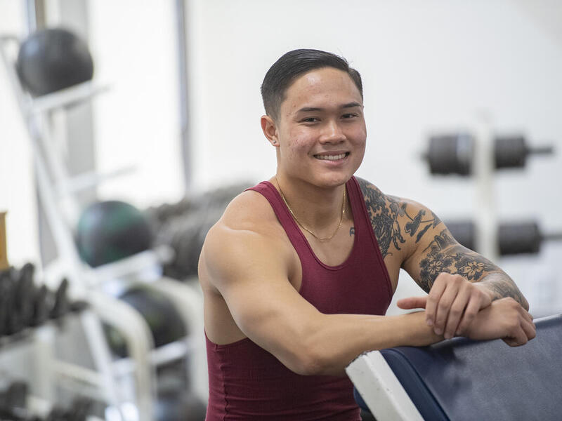 A man wearing a tank top leaning on a piece of exercising equipment. 