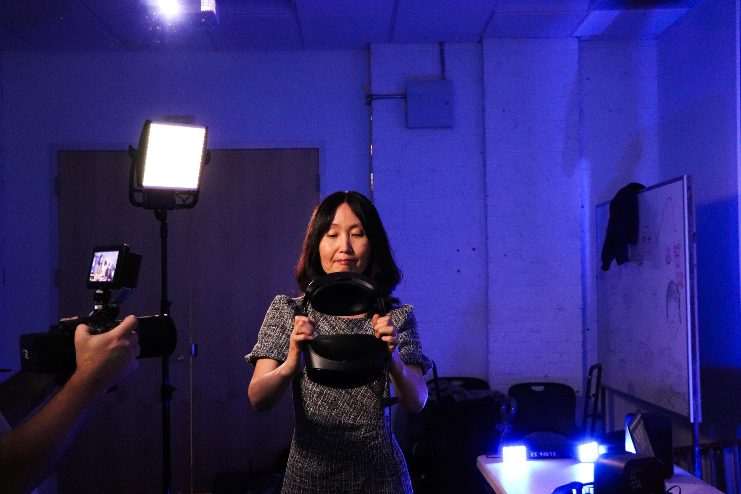 A photo of hands holding a camera that is facing a woman. The woman is holding a virtual reality headset. 