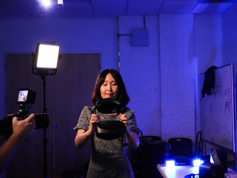 A photo of hands holding a camera that is facing a woman. The woman is holding a virtual reality headset. 