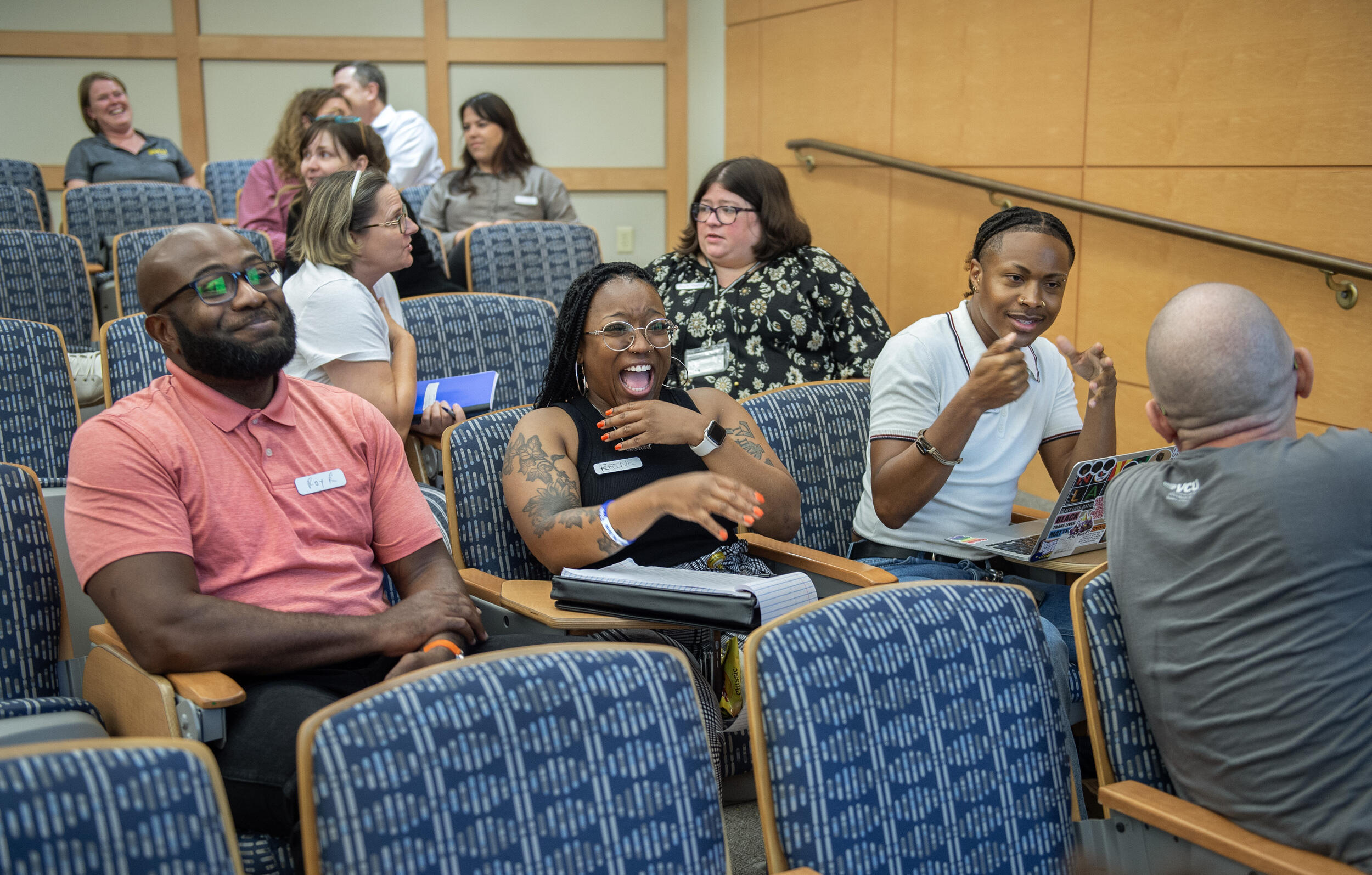 A photo of 11 people sitting in six rows of chairs. Multiple people are leaning over their charis to talk to eachother and are laughing. 