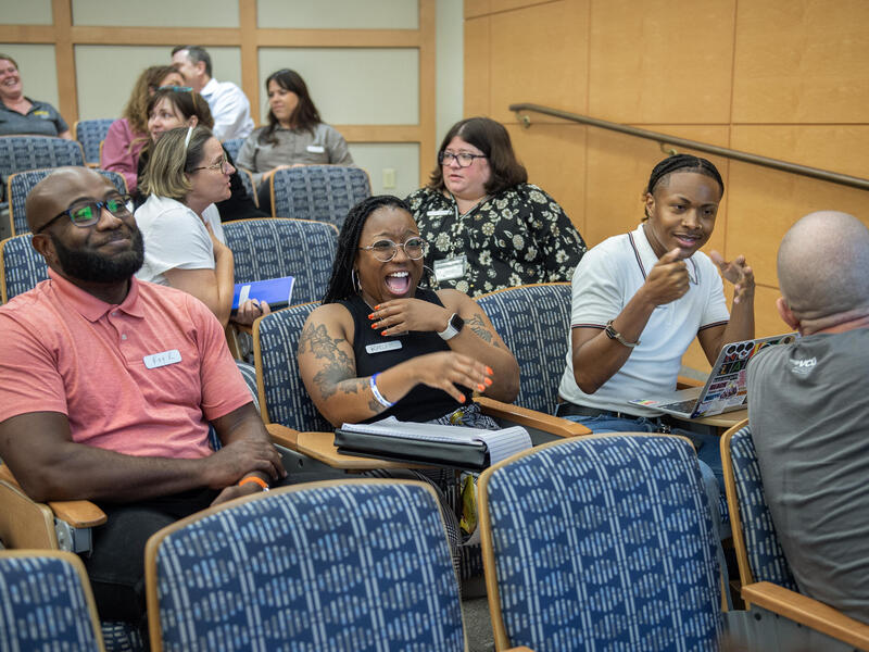 A photo of 11 people sitting in six rows of chairs. Multiple people are leaning over their charis to talk to eachother and are laughing. 